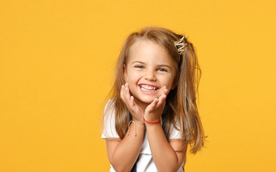 A little girl holding her face and smiling while wearing a headband with a tiara on it